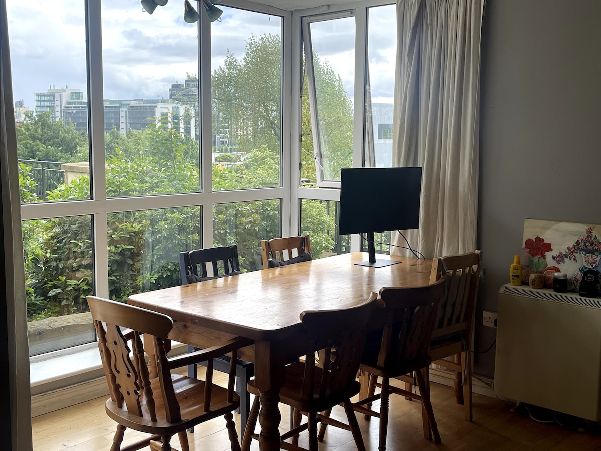 A dining area is highlighted by a wooden table surrounded by six chairs. A television is positioned on the table, with large windows providing a view of greenery outside. Bright natural light fills the space, enhancing the inviting atmosphere.