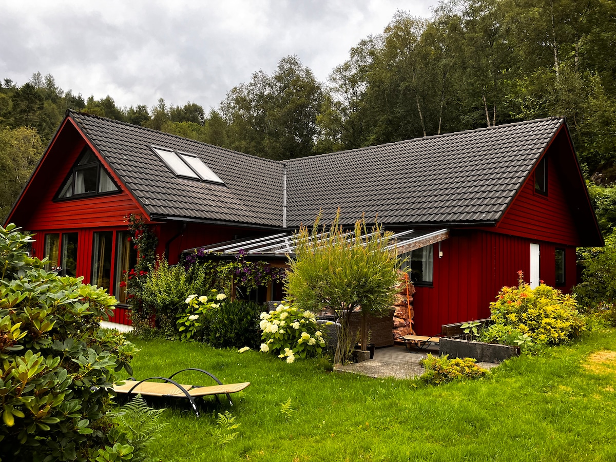 A red house with a sloped roof sits amid green grass and shrubs. Large windows allow for natural light, while a covered patio features outdoor seating. Lush greenery and varied plants surround the home, providing a serene and natural environment.