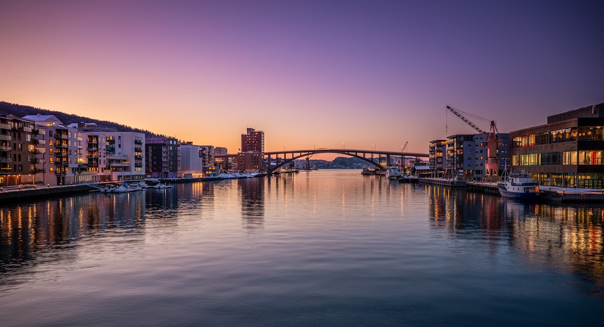 A serene view of the waterfront captures a tranquil body of water reflecting the soft hues of sunset. Modern buildings line the shore, and a bridge connects both sides, all under a gradient sky transitioning from warm orange to deep purple.