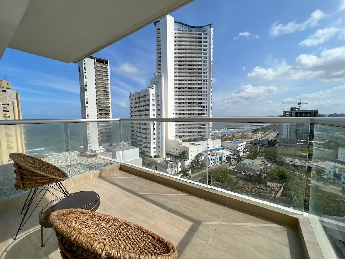 A balcony with a glass railing features two woven chairs and a small table. The view showcases nearby high-rise buildings and a direct perspective of the sea in the background. The space is brightened by natural light and open sky.