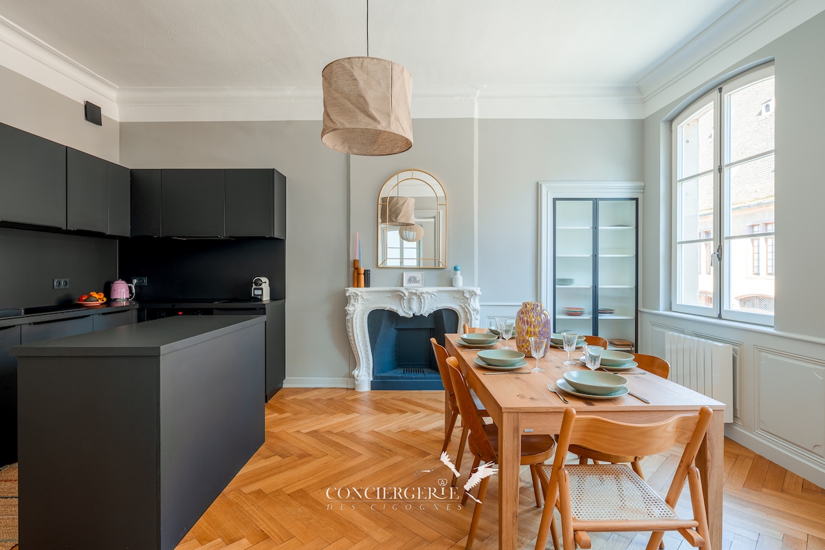 A modern kitchen and dining area feature sleek black cabinetry and a large island. A wooden dining table is set with dishes, surrounded by elegant chairs. Natural light from the large window illuminates the space, highlighting the classic fireplace and decorative mirror.