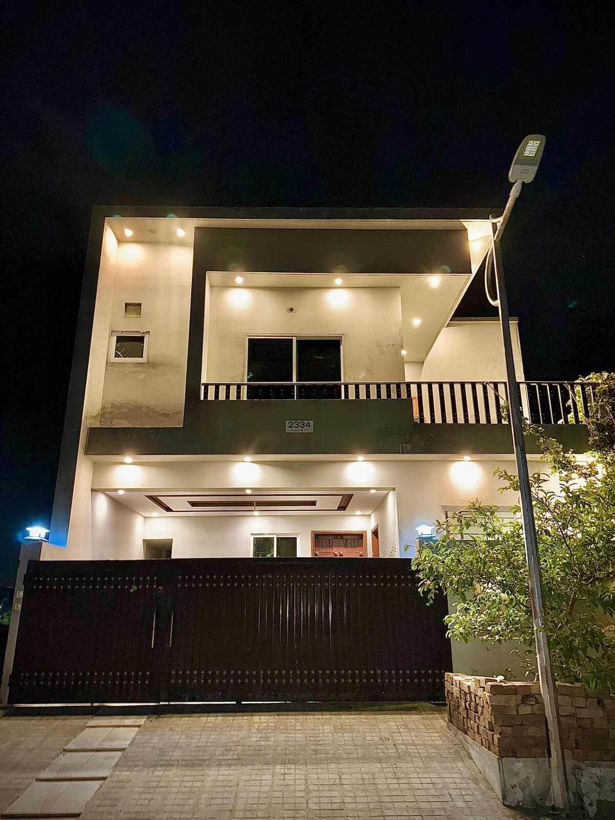 The modern exterior of the property is illuminated by strategically placed lighting, highlighting the architectural features. A welcoming front gate leads to a spacious entrance, framed by a lush garden and a smooth, paved pathway. The balcony showcases a view of the surroundings.