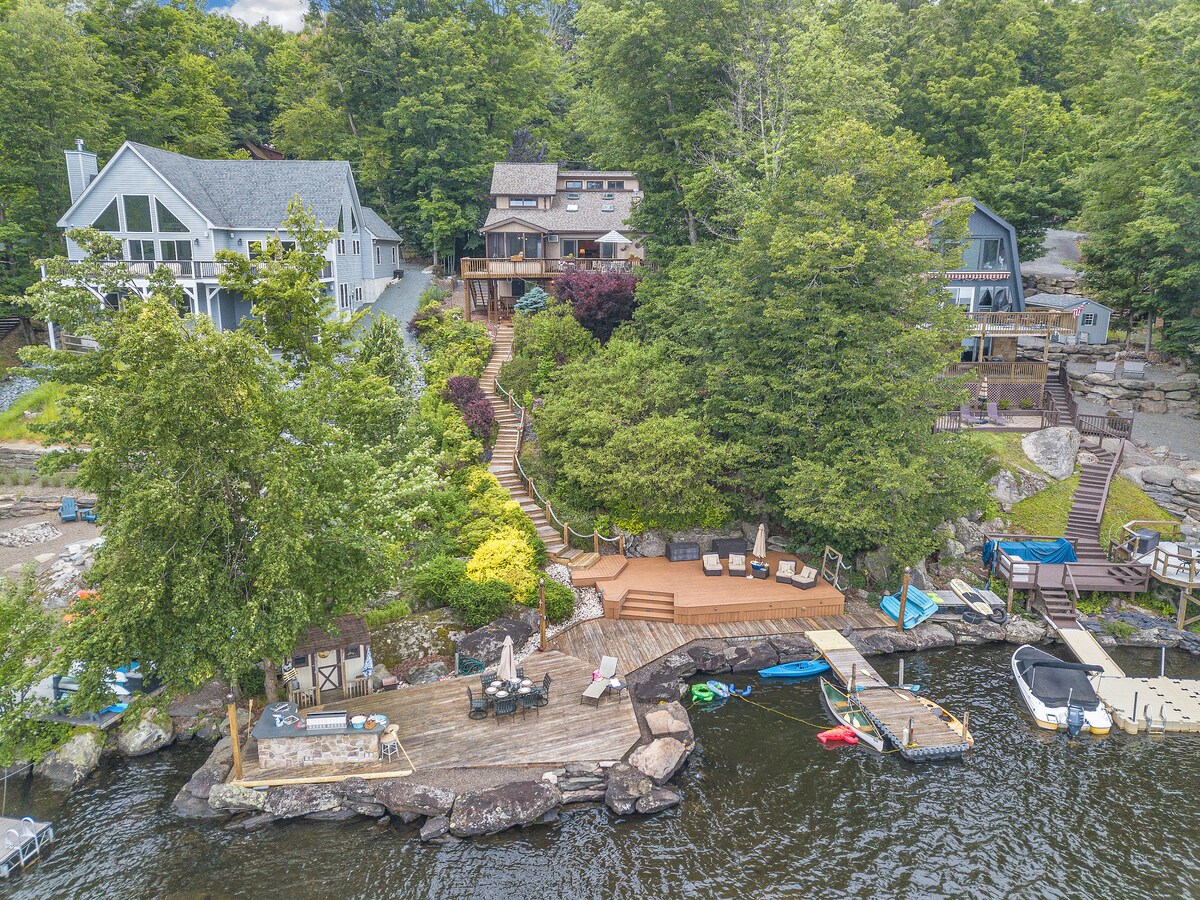 An aerial view of a lakefront property showcases a multi-level landscape with lush greenery and wooden decks. Several lounge chairs are positioned near the water, alongside a private dock equipped with kayaks and a swimming area. Surrounding homes are visible on the hillside.