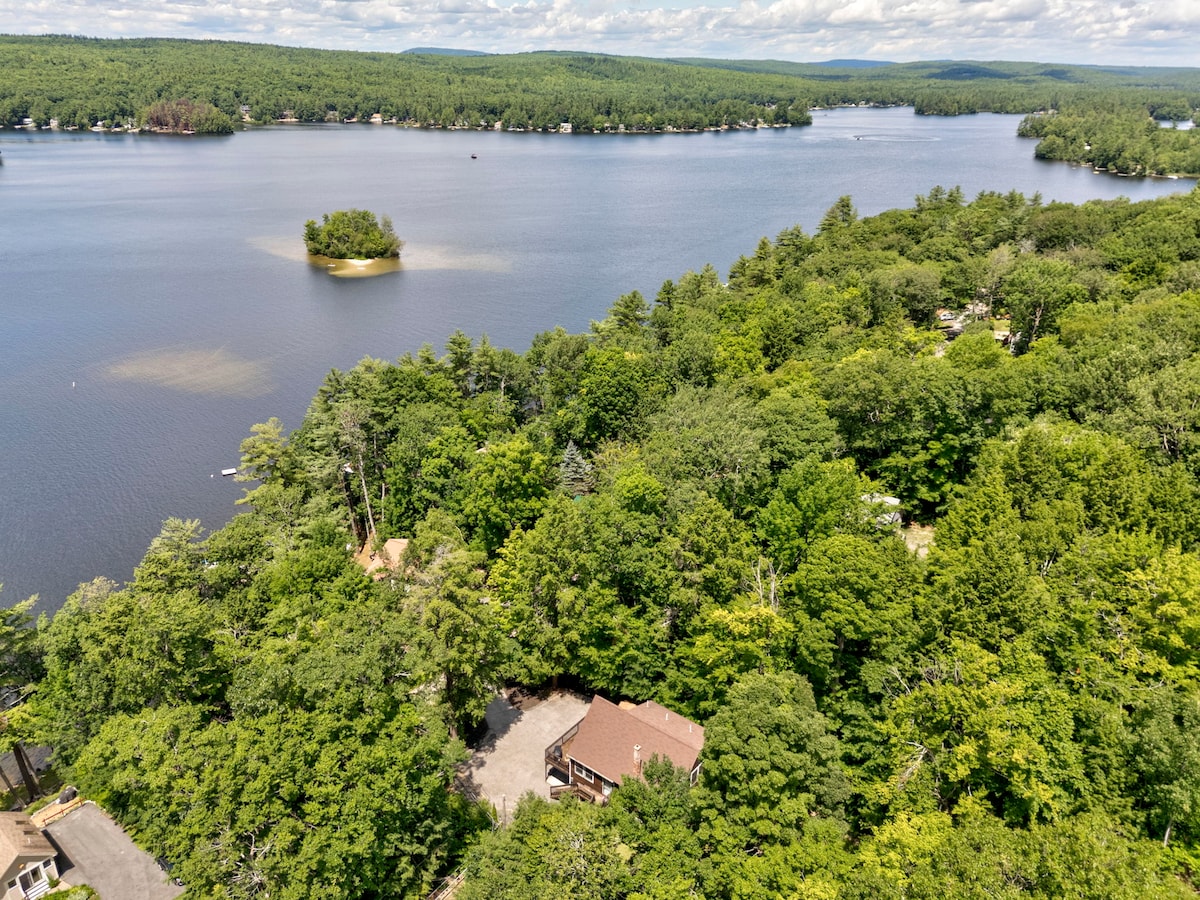 An aerial view captures the cabin nestled among lush greenery, surrounded by the calming waters of Little Sebago Lake. The scene features a small island in the lake, with various shades of blue and green reflecting a serene natural landscape.