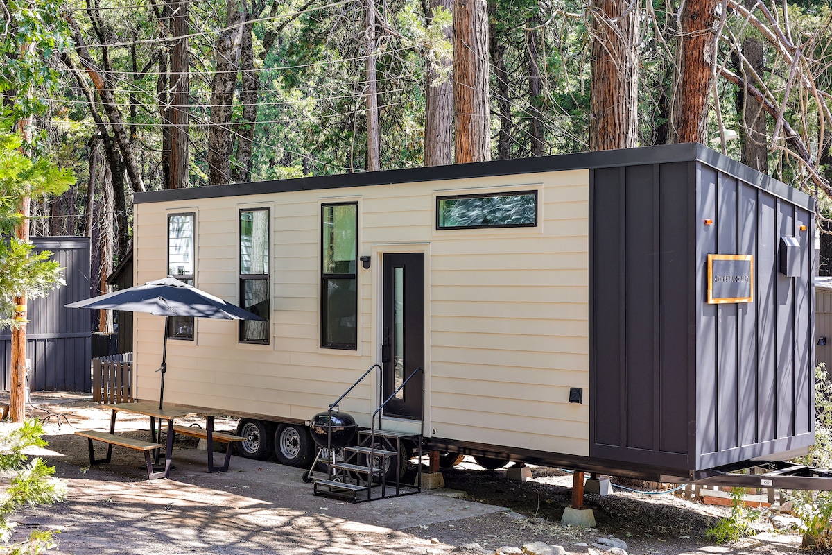 A modern tiny home is set among tall trees, featuring a light-colored exterior with dark accents. Windows provide natural light, and a small patio area is furnished with a table and an umbrella for shade.