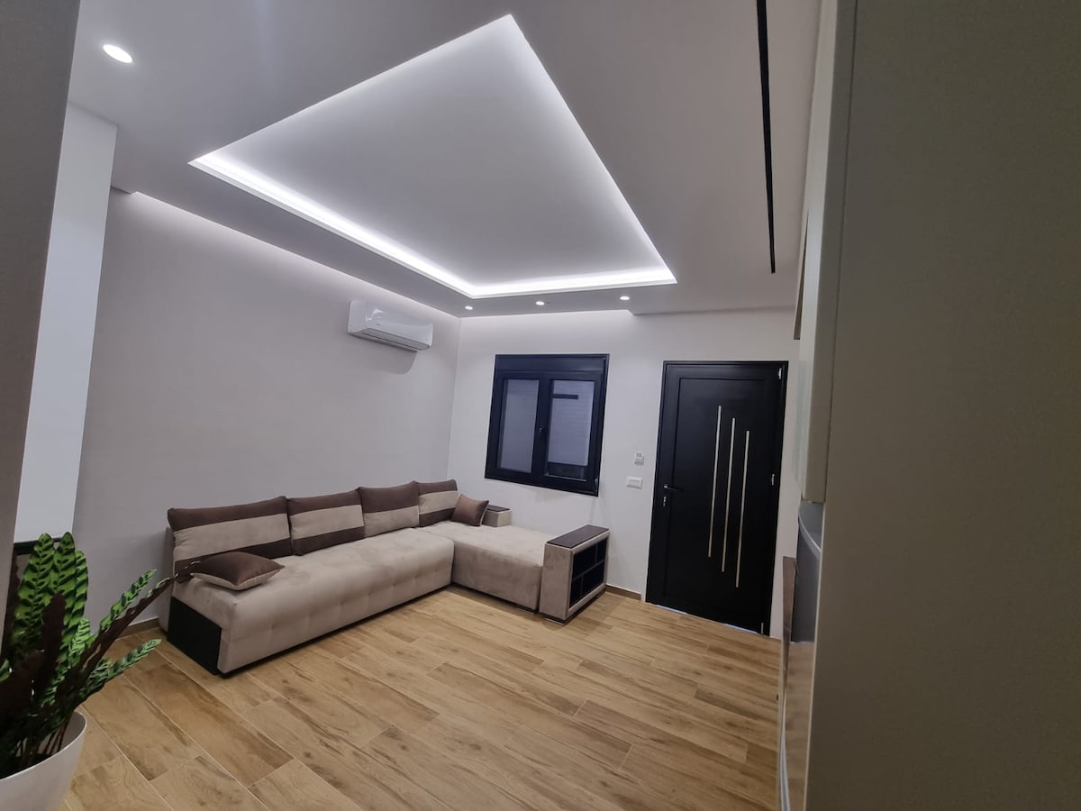 A spacious living area features a large sectional sofa arranged against a wall, complemented by a sleek coffee table. Soft, recessed lighting highlights the smooth ceiling, while a window offers a view of the outside. Light wood flooring adds warmth to the space.