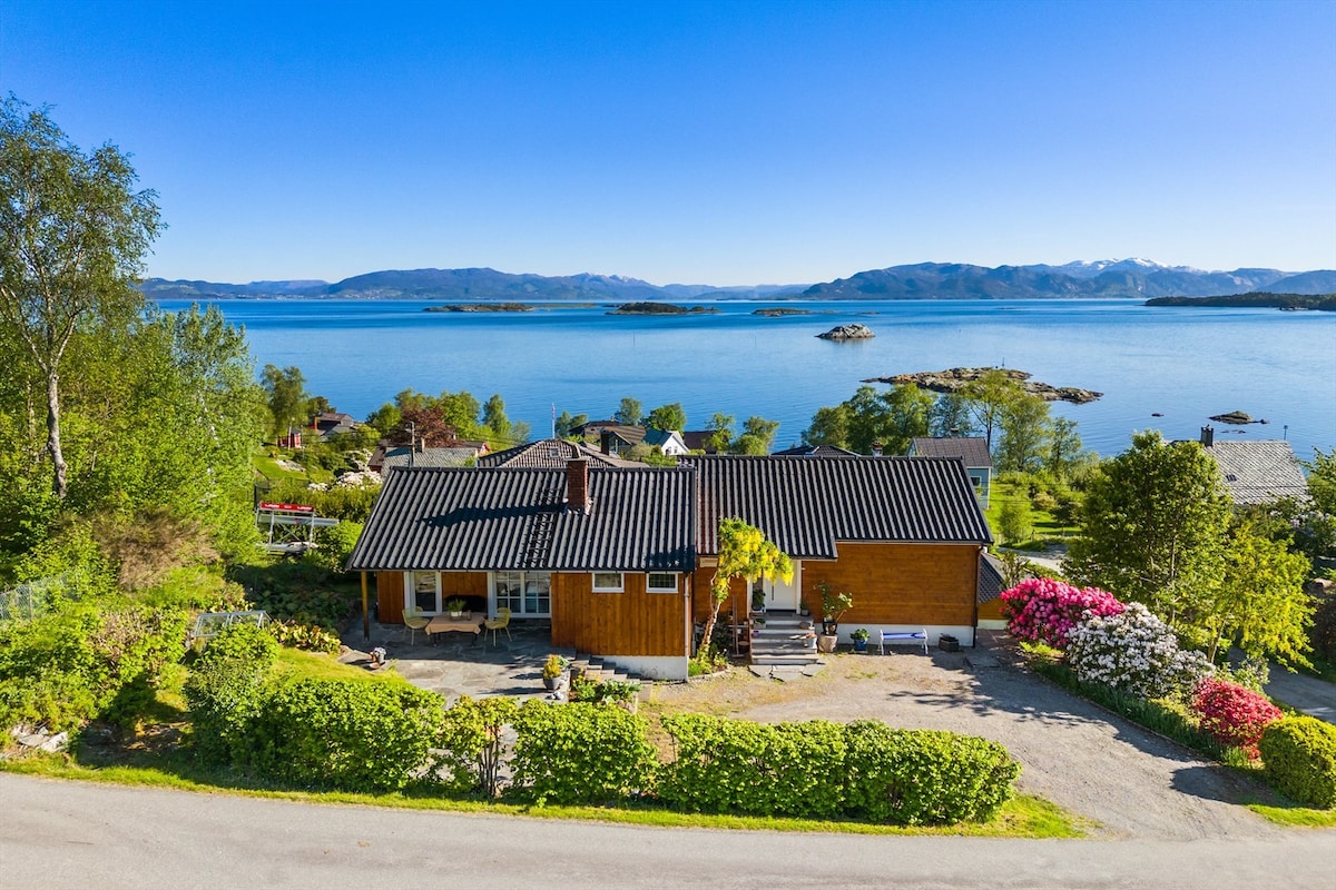 A spacious vacation home is situated by the water, offering panoramic views of the fjord. The well-maintained exterior features landscaping with vibrant greenery and blooming flowers, complemented by a gravel pathway leading to the entrance. The surrounding natural scenery is highlighted in the background.