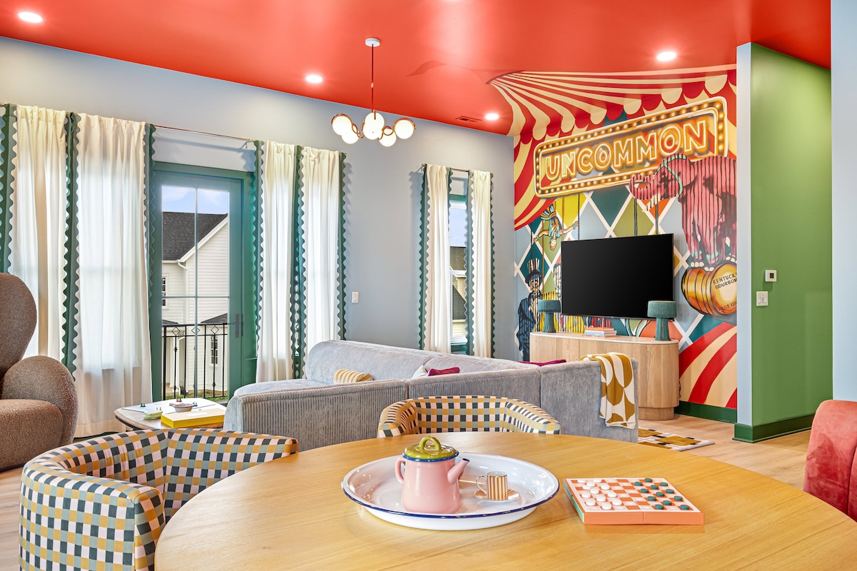 A colorful living area features a striking red ceiling and vibrant wall art depicting a circus theme. Seating includes a gray sofa, a round dining table, and unique patterned chairs. Large windows allow natural light to fill the space, enhancing the playful decor.