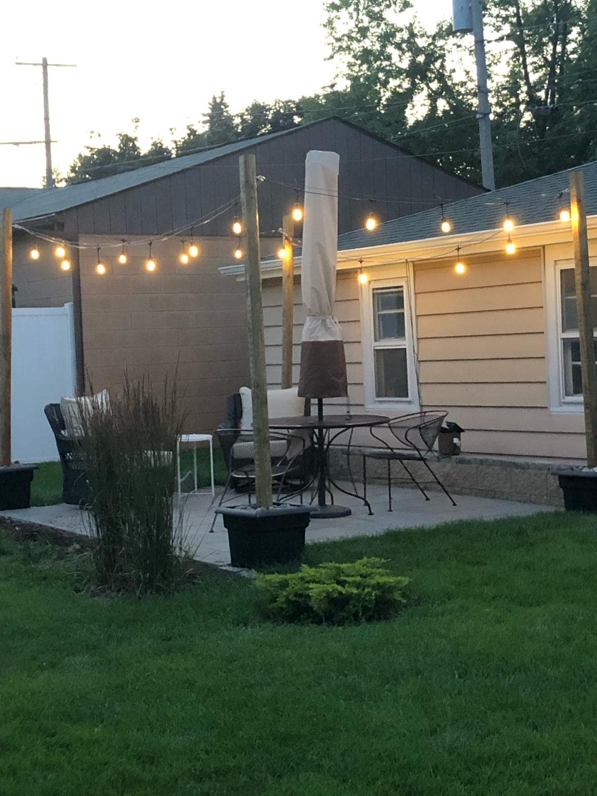 A serene backyard area is highlighted by a patio featuring a round table surrounded by several chairs. Soft lighting from string lights creates a cozy ambiance. Decorative planters with tall grass and small shrubs add a touch of greenery to the space.