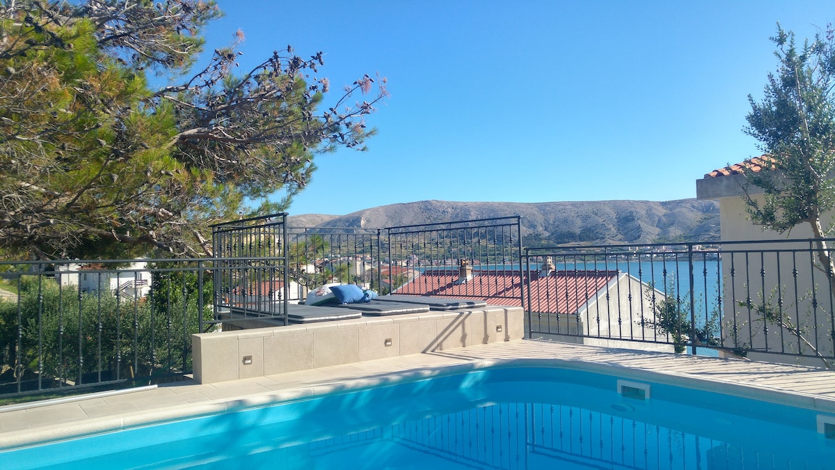 A pool area is shown overlooking a scenic view of the landscape and rooftops. The crystal-clear water of the pool reflects the blue sky. Sun loungers are positioned nearby for relaxation, framed by lush greenery and enhancing the overall serene setting.