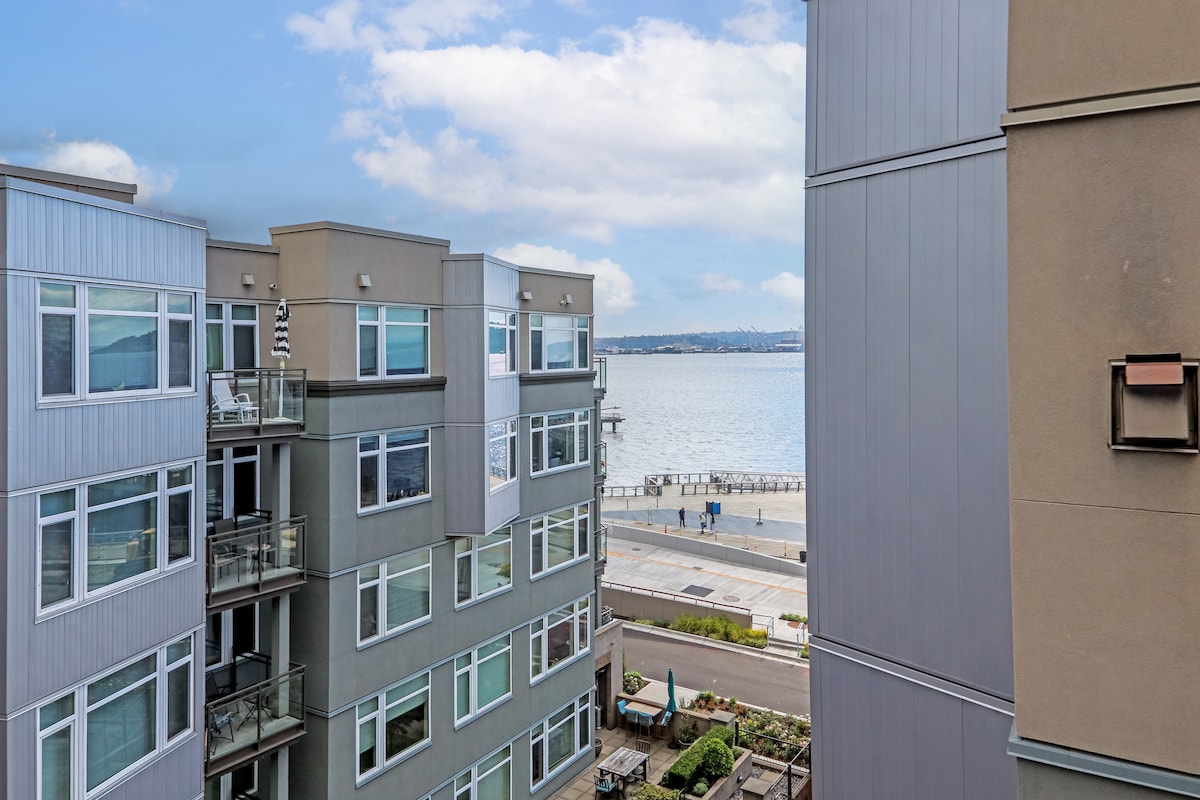 A view showcases the waterfront beyond the neighboring buildings, revealing Elliott Bay and a nearby promenade. The surrounding apartments exhibit modern architectural design, with large windows that invite natural light into living spaces.