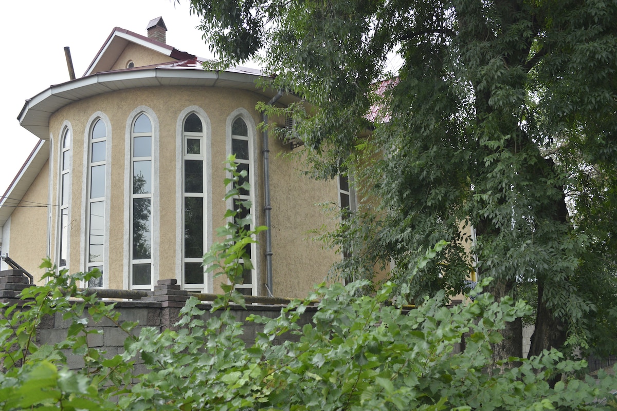 A round structure features multiple tall, arched windows that allow natural light to fill the interior. The exterior is painted in a soft yellow tone, surrounded by lush green foliage. A red roof adds a contrasting touch to the overall design.