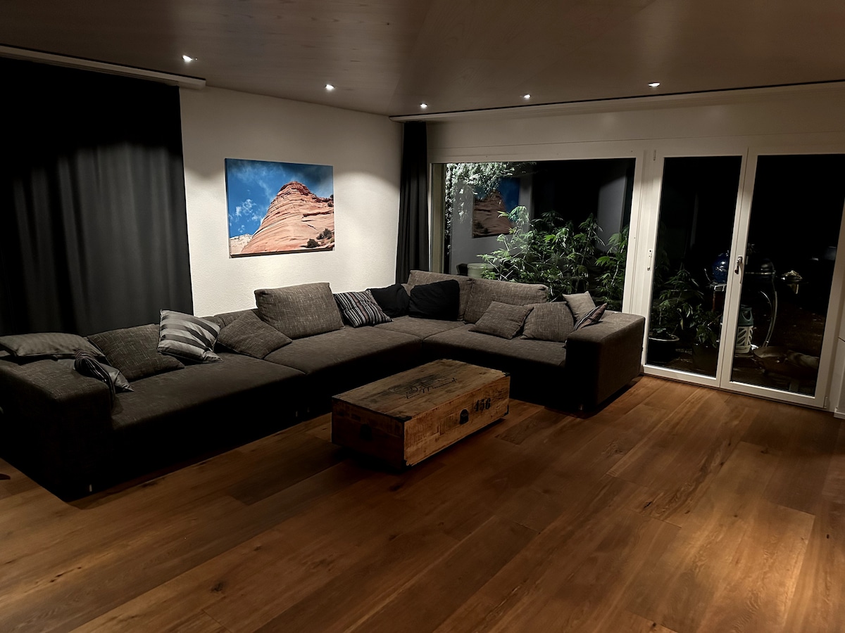 A spacious living area features a large sectional sofa adorned with decorative cushions. A wooden coffee table sits in front, while an artwork depicting rocky terrain is displayed on the wall. Large glass doors offer access to an outdoor space, surrounded by greenery.