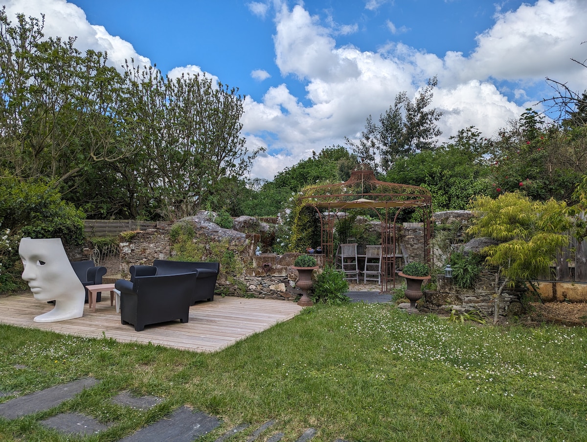 A serene outdoor space features a wooden deck with two comfortable black chairs and a small table. A vine-covered structure offers shade, surrounded by lush greenery and flowering plants. The garden's rustic stone walls add character to the peaceful setting.