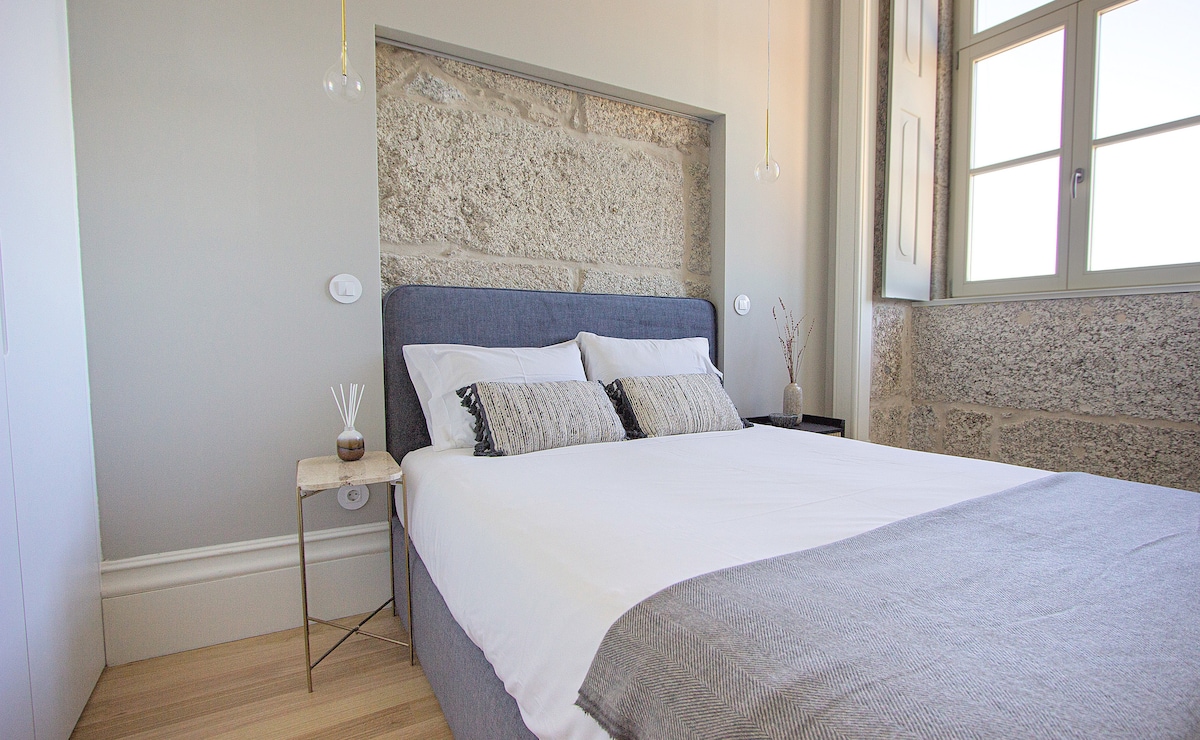 A comfortable bedroom features a spacious bed with a light grey throw and patterned pillows. Stone walls are visible, adding a textured look. A small bedside table holds decorative items, while a window with multiple panes allows natural light to illuminate the space.