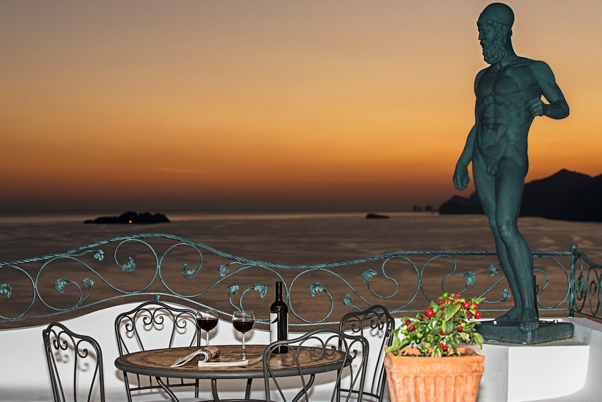 A terrace overlooking the sea is adorned with a statue, wrought-iron chairs, and a wooden table set for two. Two glasses of red wine sit alongside a bottle, accompanied by a potted plant. A stunning sunset casts warm tones over the water.