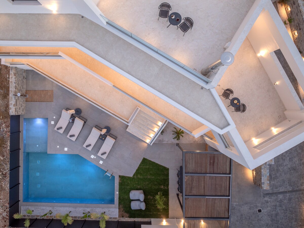 An aerial view showcases the villa's inviting outdoor space, featuring a swimming pool surrounded by premium sun loungers. Cozy dining areas with small tables are visible on the terrace, creating delightful spots for relaxation. Lush greenery and minimalist landscaping contribute to the serene ambiance.