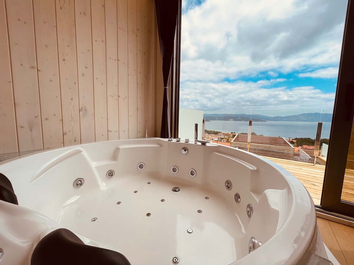 A spacious jacuzzi is positioned near large windows, offering panoramic views of the landscape and water beyond. The surrounding walls are finished in light wood, creating a serene atmosphere.