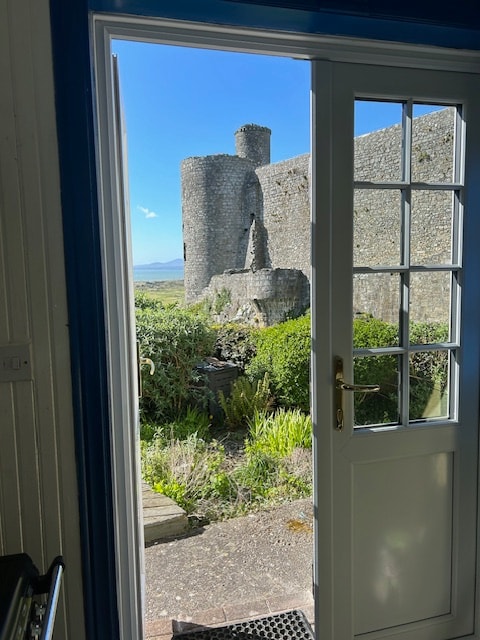 A view is presented from an open door, showcasing a stone wall and turret of Harlech Castle in the background. Lush green foliage surrounds the entrance, with a pathway leading into the garden. Bright blue skies enhance the inviting atmosphere.