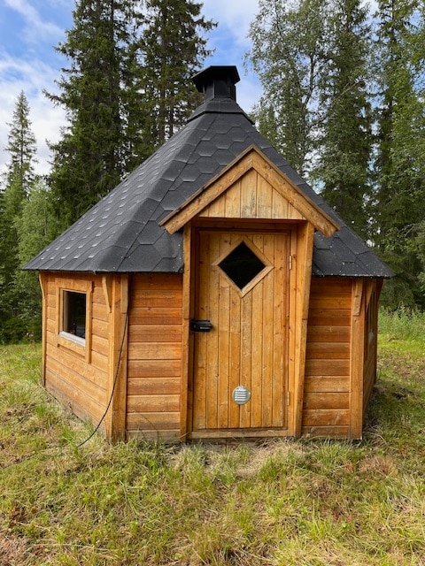 A wooden cabin with a triangular roof and a chimney is situated among tall trees. The front door features a diamond-shaped window, while smaller windows are located on the sides. The cabin is surrounded by grassy ground and a natural forest backdrop.