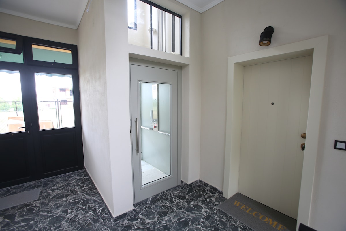 A well-lit entryway is presented with polished marble flooring. A modern glass door provides access to the apartment. Adjacent is a solid wooden door featuring a welcome mat. Natural light filters in through a nearby window, enhancing the spaciousness of the area.
