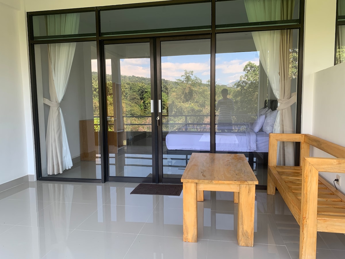 The image shows a bright and airy space with floor-to-ceiling glass doors that open to a balcony overlooking lush greenery. A wooden table and seating area are positioned within the room, and a spacious bed can be seen in the background, framed by sheer curtains.