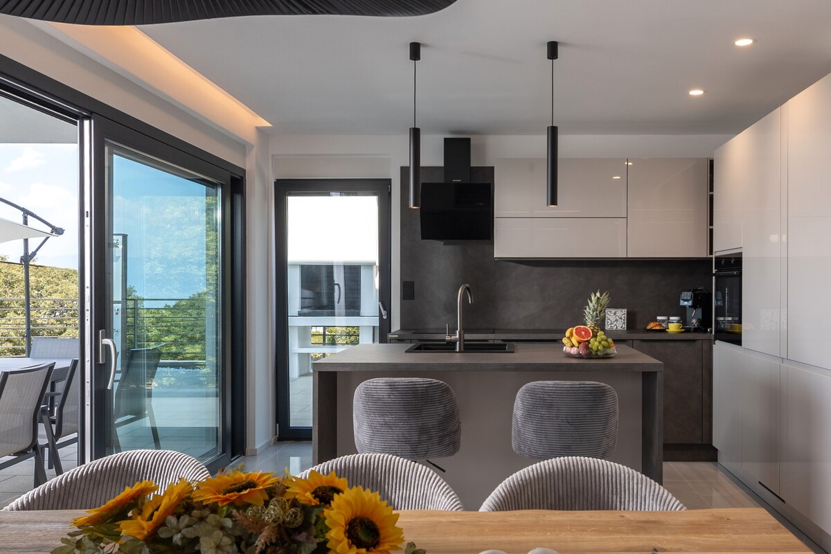 A modern kitchen is featured with sleek cabinetry and a central island equipped with a sink. Large glass doors open to a balcony, allowing natural light to fill the space. A dining table adorned with a floral centerpiece complements the inviting atmosphere.