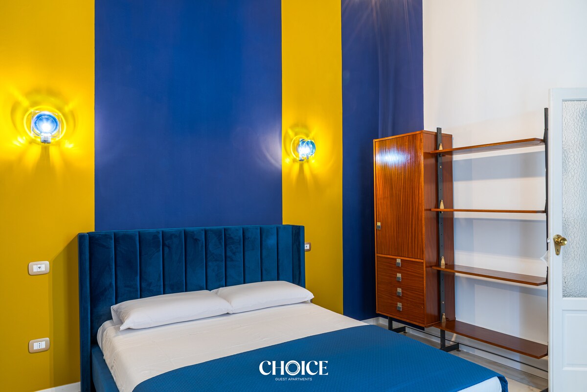 A cozy bedroom features a blue and yellow accent wall with a spacious bed dressed in white linens. Elegant wall sconces provide soft lighting, while a wooden shelf stands nearby, offering a blend of storage and style.