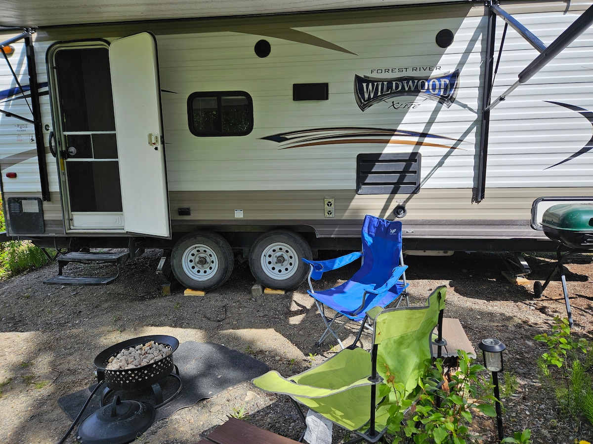 The exterior of a travel trailer is shown, featuring a door leading inside. Two outdoor chairs are placed nearby, along with a small fire pit positioned on gravel. Soft lighting is provided by a few solar garden lanterns, enhancing the outdoor space.