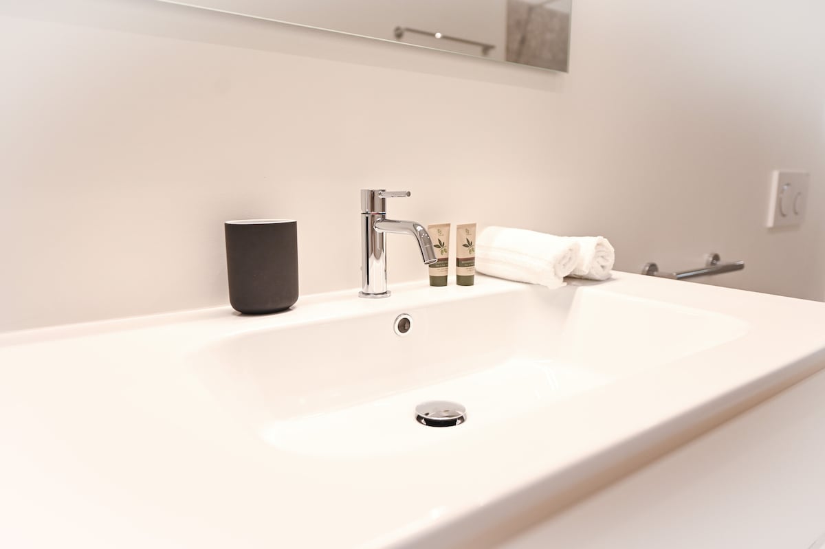 A clean and modern bathroom sink area is presented, featuring a sleek faucet and a white countertop. Adjacent items include a dark tumbler, two small toiletries, and a neatly folded white towel, contributing to a tidy and efficient space.