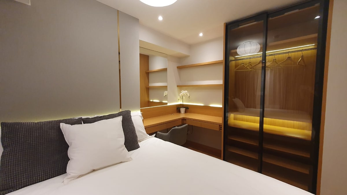 A modern bedroom features a spacious bed with neutral linens. A minimalist desk and chair are positioned against a wall of built-in shelves. A sleek glass wardrobe reveals wooden details and provides ample storage space. Soft lighting accentuates the room's contemporary design.