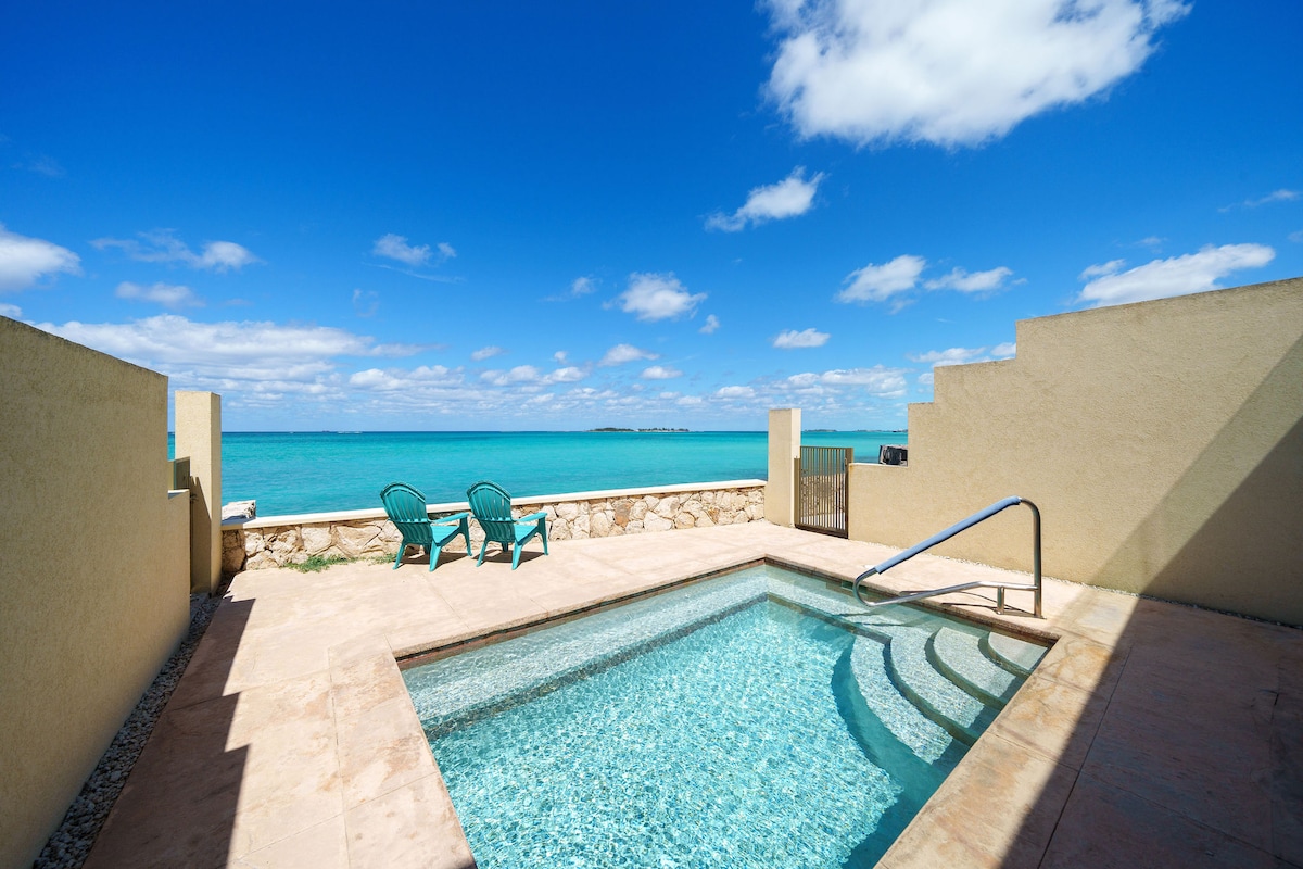 An inviting outdoor space features a small plunge pool, surrounded by a smooth stone deck. Two turquoise lounge chairs are positioned to provide views of the tranquil turquoise waters beyond. The sky above is filled with scattered clouds, enhancing the bright atmosphere.
