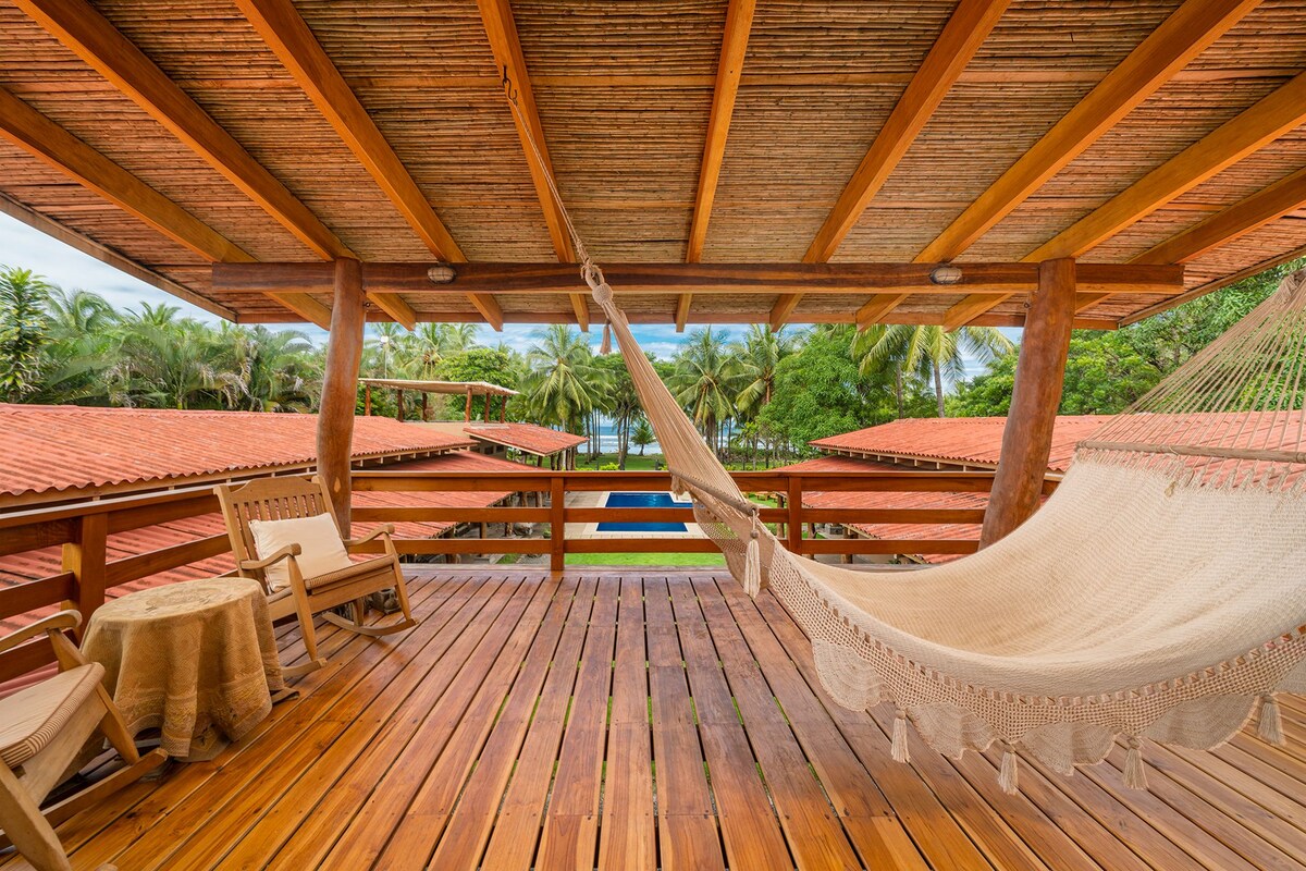 A spacious wooden deck features a woven hammock, inviting relaxation. Two wooden chairs and a small table complement the area. Below, lush greenery surrounds a serene pool, with a view of palm trees and the ocean in the distance.
