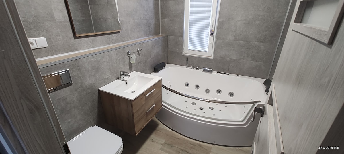 A modern bathroom features a whirlpool tub with adjustable jets and a shower. A clean white vanity with a sink sits next to a toilet. Natural light enters through a nearby window, illuminating grey tiled walls and wooden accents.