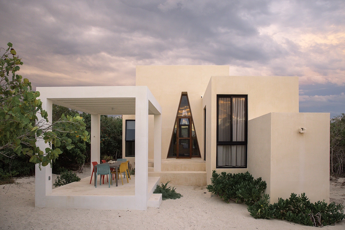 A modern coastal home is set against a cloudy sky, featuring a unique triangular window. The open terrace showcases a dining area with colorful chairs, surrounded by natural foliage and sandy ground, inviting a relaxed outdoor atmosphere.