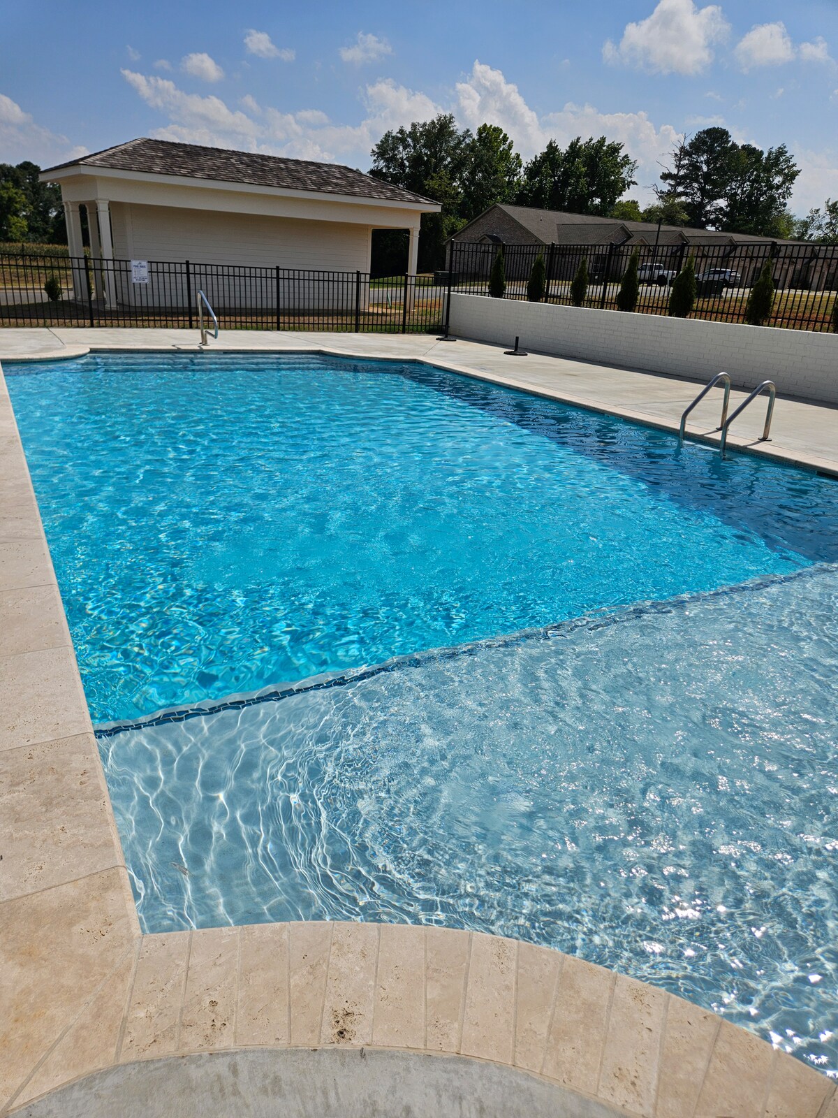 A large outdoor swimming pool glistens under the sunlight, surrounded by a clean coping and a secured fence. The pool features a shallow area on one side, with stairs leading into the water, inviting relaxation and enjoyment.