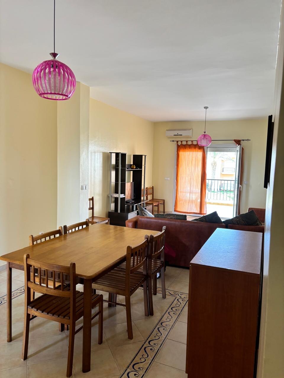 A dining area features a wooden table surrounded by six chairs, illuminated by two pink pendant lights. The space includes a comfortable seating area with a brown couch, a television unit, and access to a balcony that allows natural light to brighten the room.