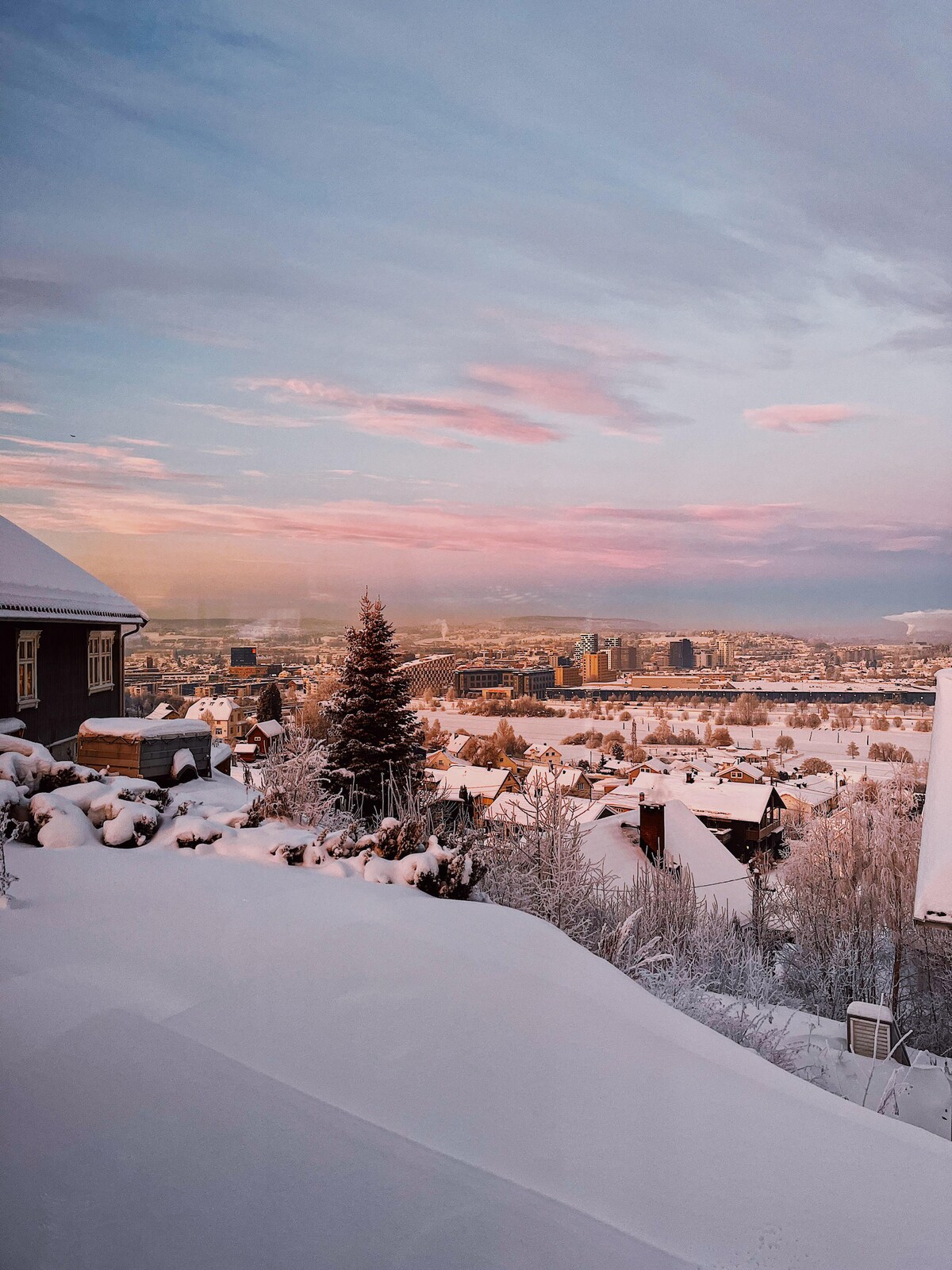 A panoramic view of a snowy landscape is captured, showcasing a city nestled in the valley below. The harmonious colors of the sky transition from soft pink to blue, while covered rooftops and snow-laden trees depict a serene winter scene.