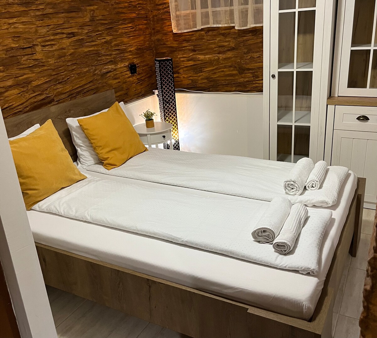 A comfortable sleeping arrangement is displayed, featuring two spacious beds positioned side by side. Each bed is neatly made with white linens and accented by yellow throw pillows. Soft textures and natural wood elements create a cozy environment.