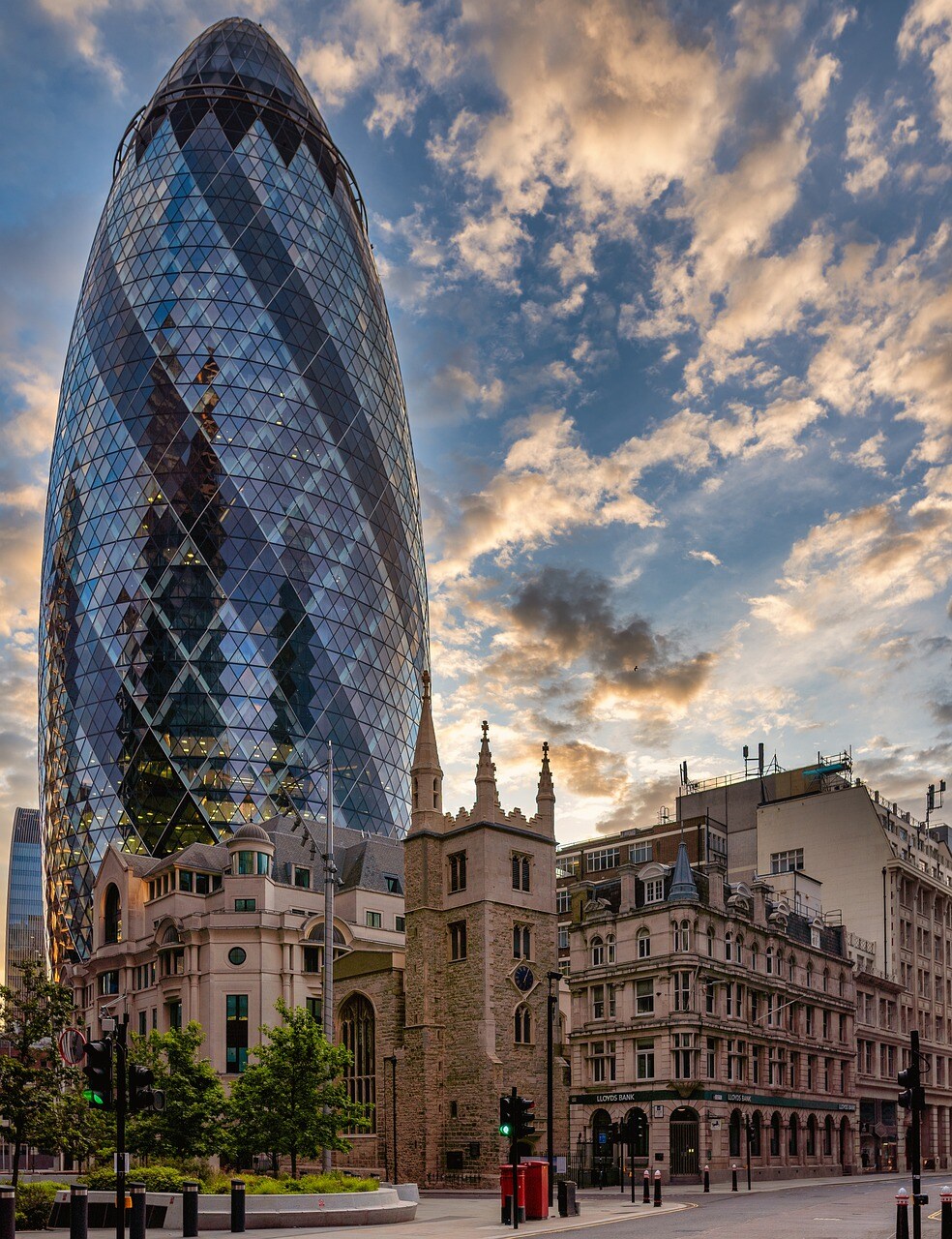 A modern skyscraper, known as the Gherkin, rises prominently among a mix of historic buildings. Its glass façade reflects the sky, while clouds are painted in hues of orange and blue. Nearby, a stone structure with pointed arches stands in contrast to the sleek design.