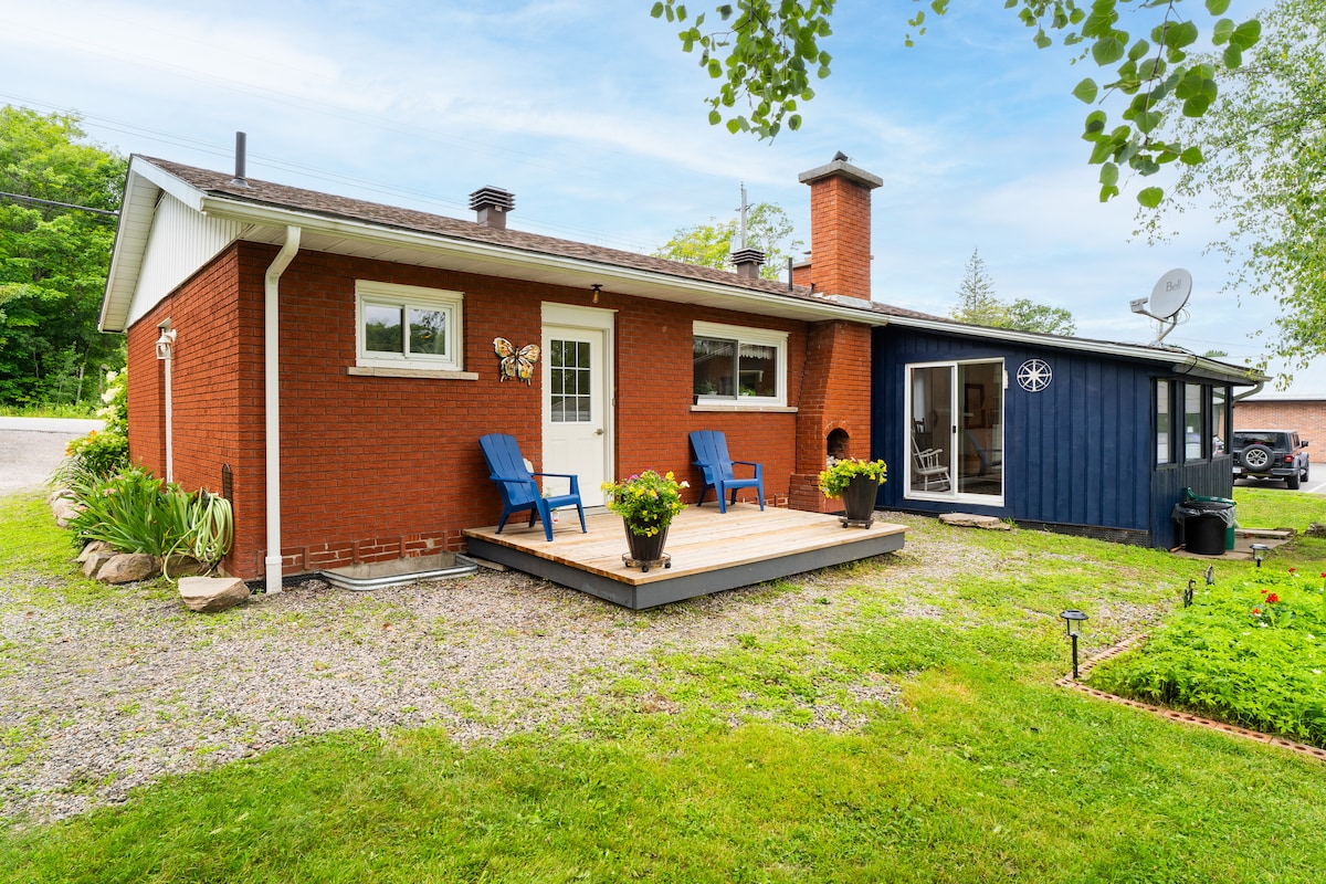 The exterior of the home features a brick facade with a small deck adorned with two blue chairs. A well-maintained lawn surrounds the property, complemented by modern outdoor decor. A cozy sunroom with glass panels is visible on one side, inviting natural light into the space.