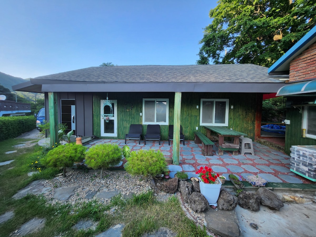 A charming exterior view of the property features a green wooden faรงade complemented by large windows. A stone pathway leads to a patio area furnished with chairs and a picnic table. Potted plants and small shrubs are arranged for a welcoming touch.