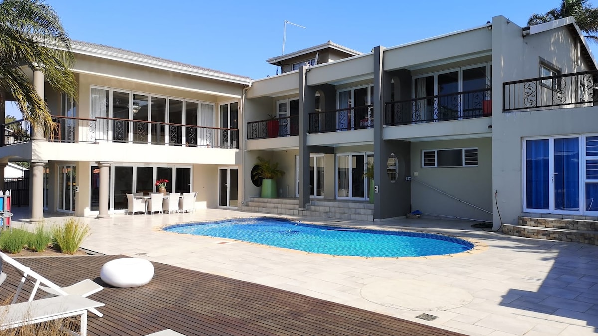 The outdoor area features a sparkling pool surrounded by a spacious patio, complemented by lounge chairs and a dining set. Two levels of the home are visible, with large windows providing ample natural light and views of the pool area.