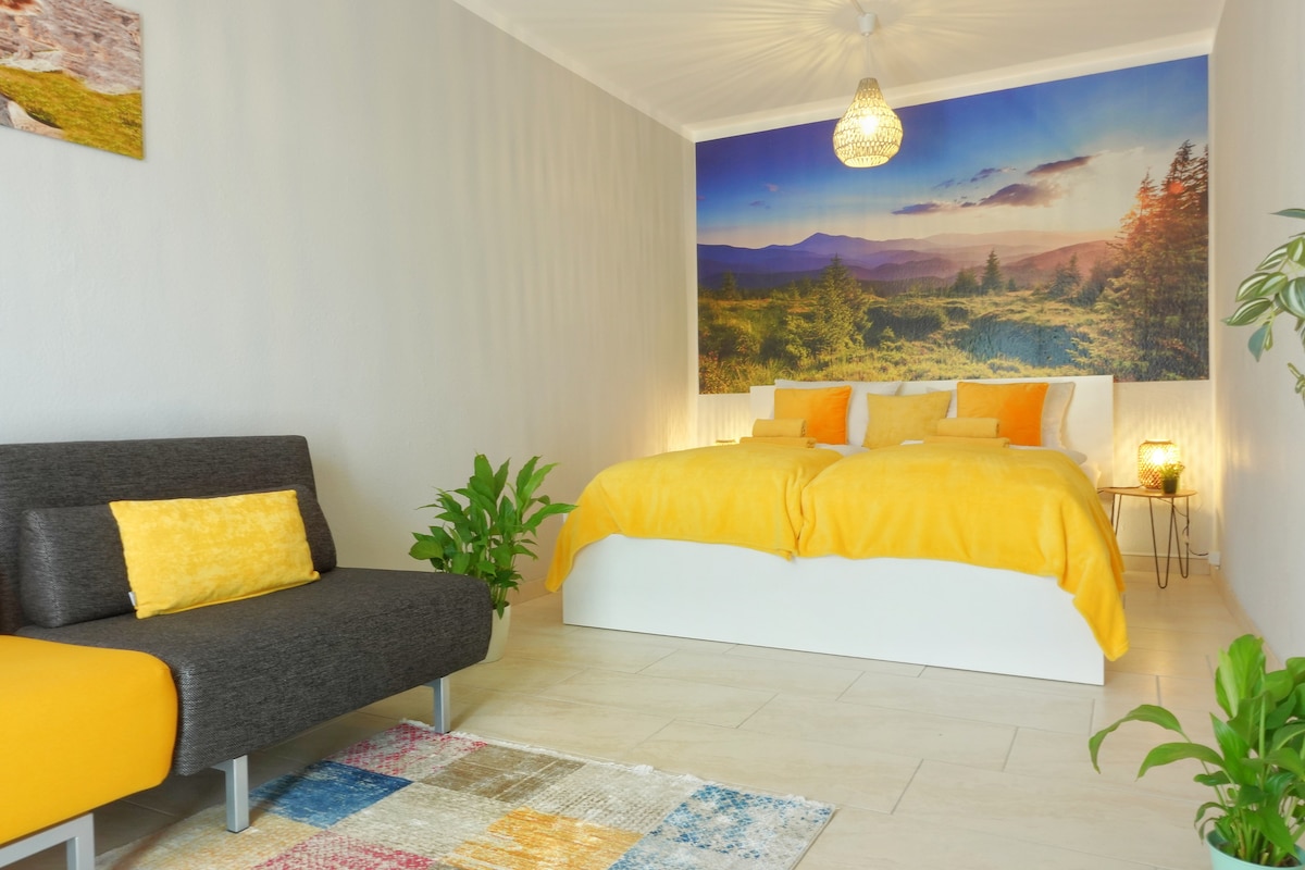 A bright bedroom features a large mural depicting a scenic landscape. A spacious bed with yellow linens is paired with decorative pillows. A cozy seating area is complemented by a gray sofa, while potted plants and a colorful area rug enhance the inviting space.