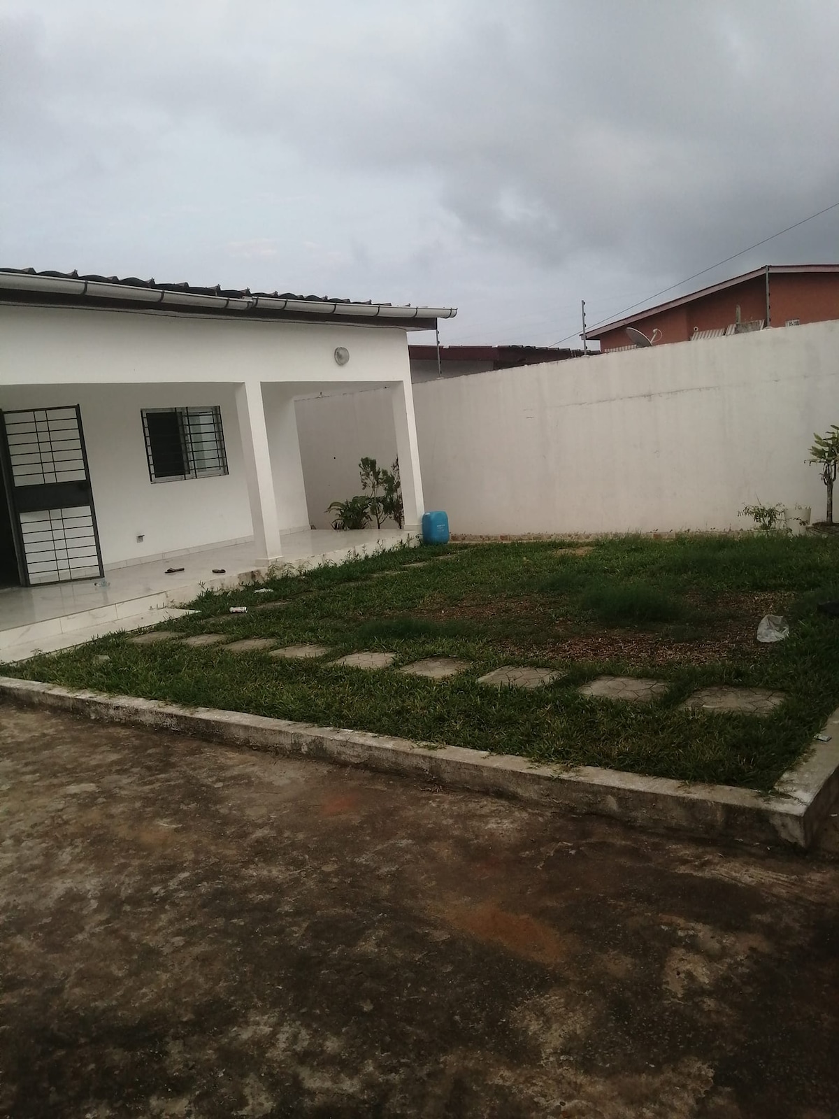 A spacious outdoor area is shown, featuring a small garden with uneven stone pathways leading through green grass. A white wall encloses the space, with a light-colored building partially visible. A blue water container is positioned near the garden.
