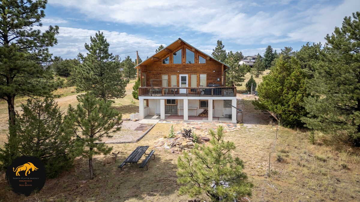 The cabin is showcased from an aerial perspective, highlighting its wooden structure complemented by a second-story balcony. Surrounding pine trees create a natural setting, with a spacious patio area featuring a picnic table. The expansive landscape emphasizes the cabin's serene hillside location.