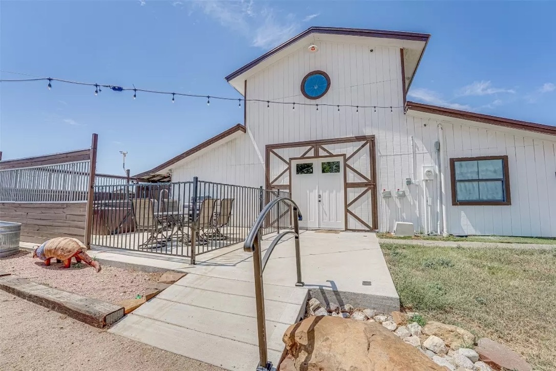 The exterior of the farmhouse features a spacious entryway with a ramp, providing accessibility. A patio area with outdoor seating is visible, alongside decorative stones and a charming turtle sculpture. String lights are hung above, adding a welcoming touch to the outdoor space.