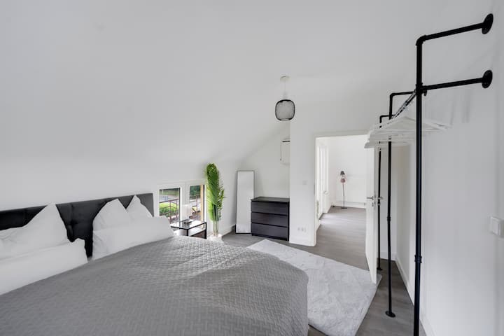 Тази спалня съчетава комфорт и стил с познатото пружинно легло, елегантни нощни шкафчета и настолни лампи, както и отворен гардероб. Достъпът до всекидневната на заден план създава отворена и приветлива атмосфера