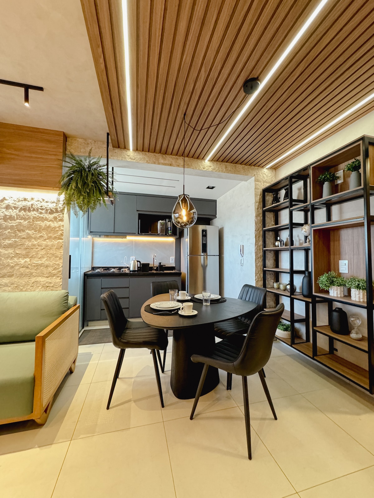 A modern dining area features a round black table surrounded by four sleek black chairs. Above, a pendant light illuminates the space. The open kitchen includes dark cabinetry and a silver refrigerator, while a green plant and decorative shelf provide visual interest.