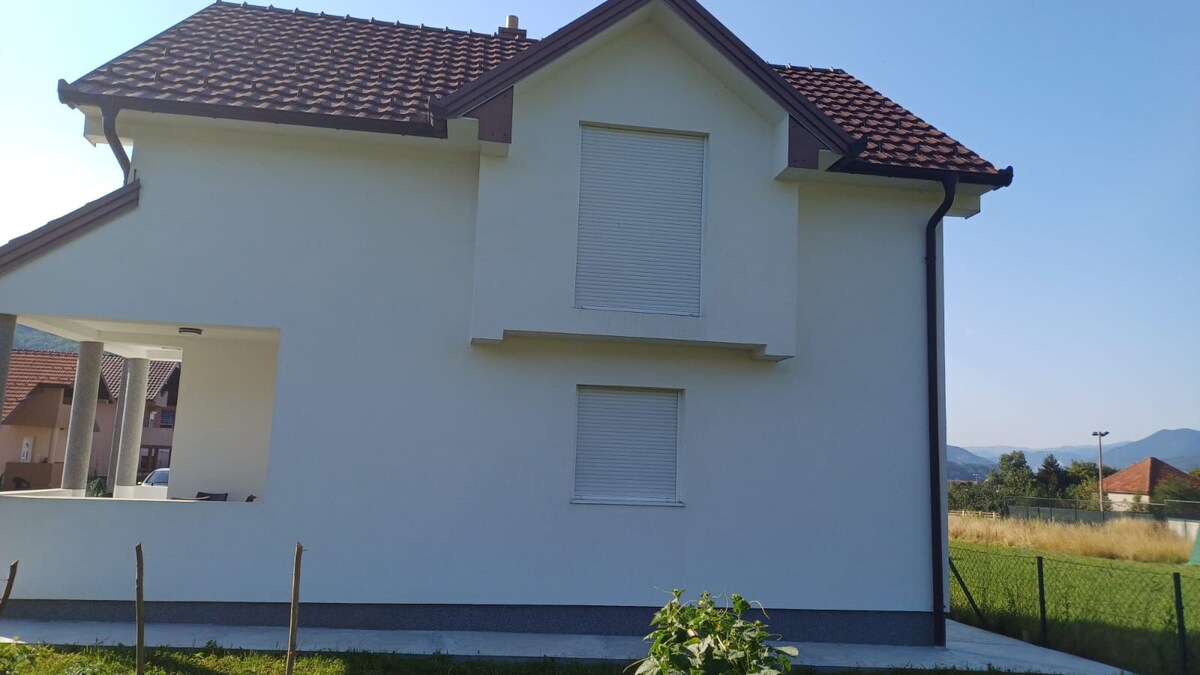 A two-story house displays a clean white facade under a clear blue sky. The roof is finished with brown tiles, and covered windows suggest a peaceful environment. A grassy area surrounds the home, enhancing its tranquil setting.